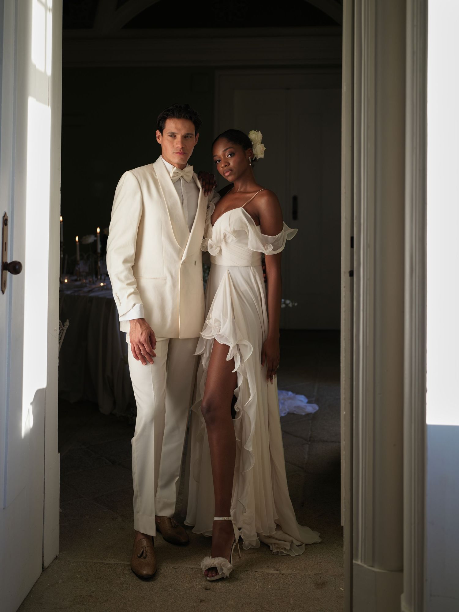 A couple in elegant all-white attire poses together in a hallway with multiple doorways.