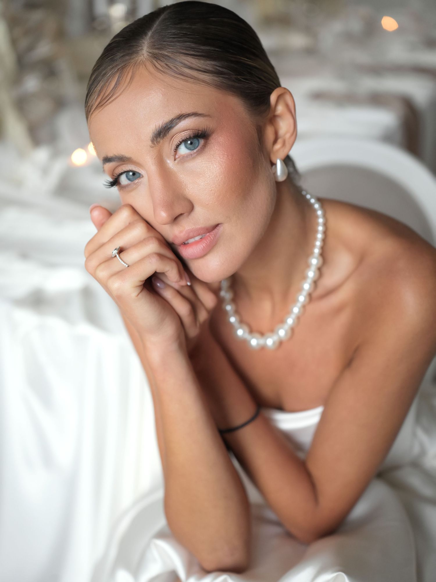 Elegant woman wearing pearl necklace and earrings in white dress, posing gracefully with hands near face.
