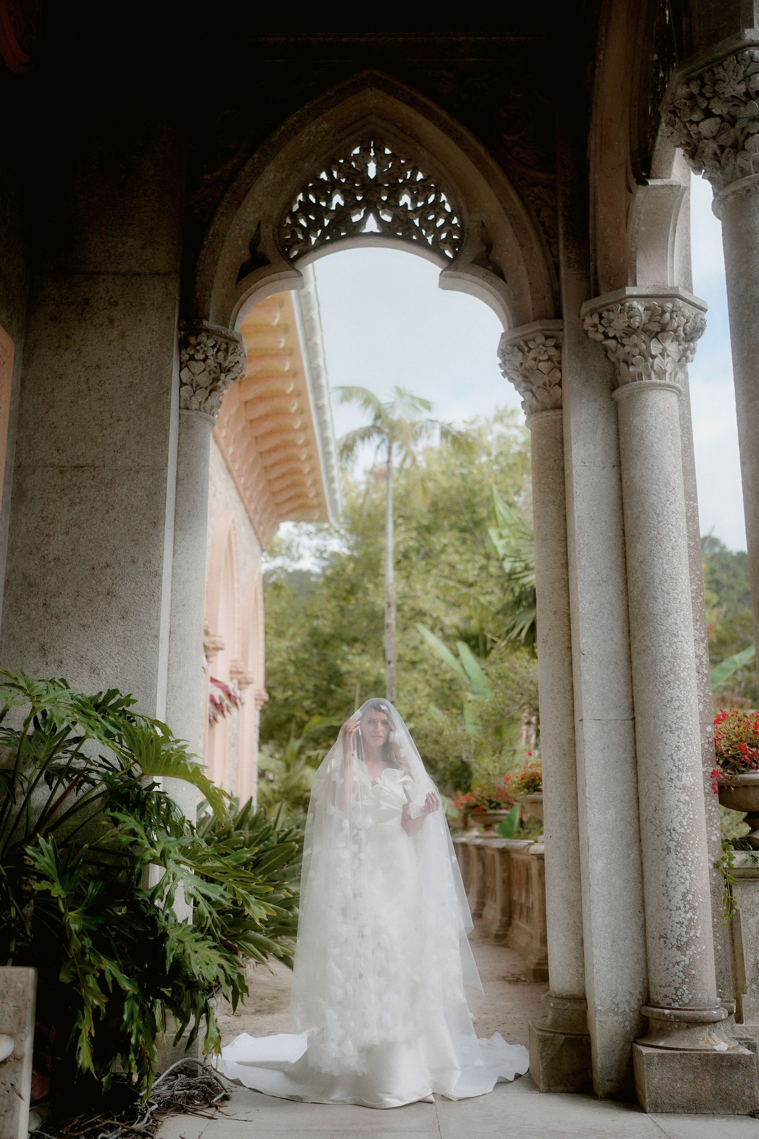 A bride in a flowing white wedding gown stands elegantly beneath ornate stone archways surrounded by lush tropical foliage.