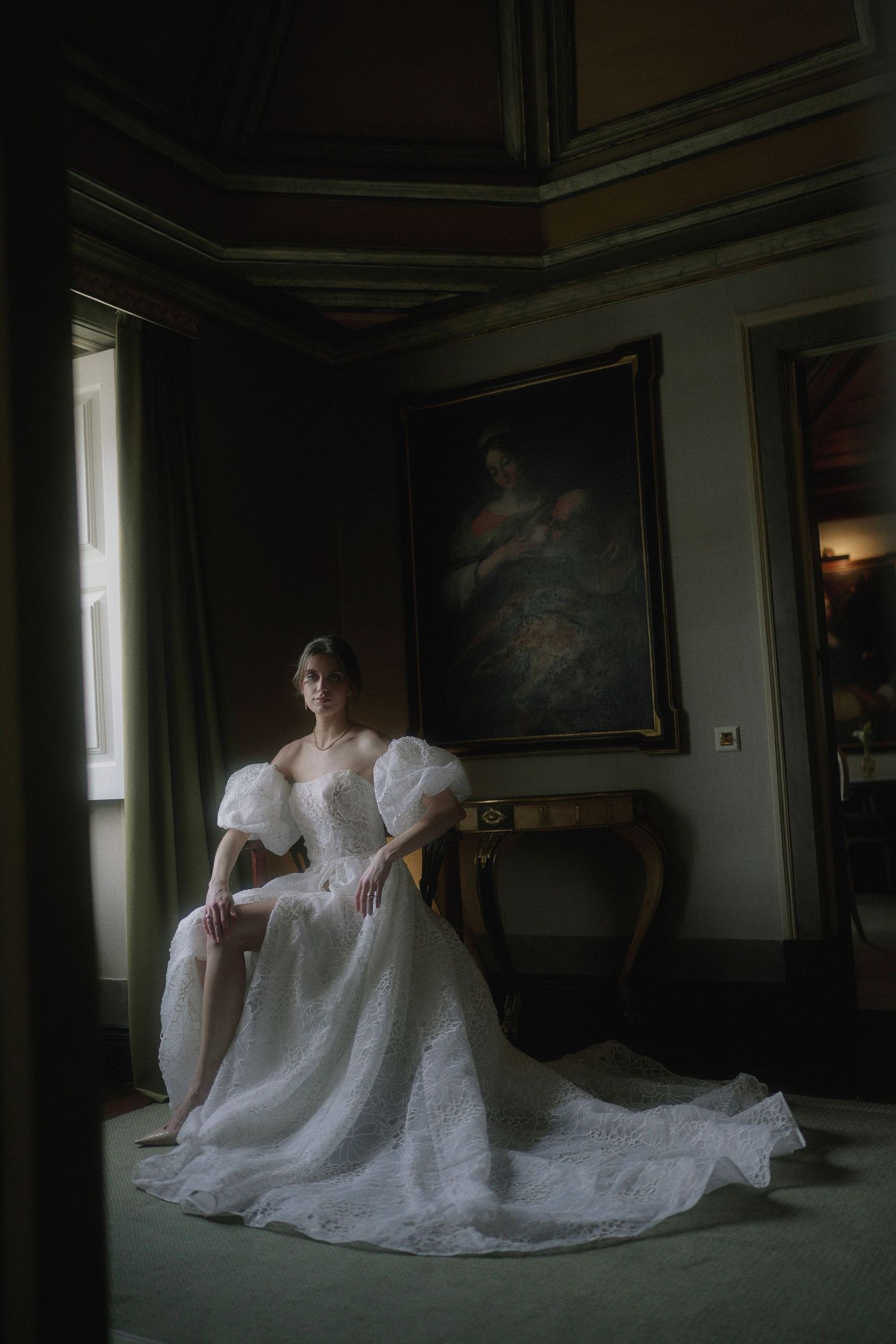 A figure in a white lace wedding dress reclines on a dark floor in a dimly lit room with windows.