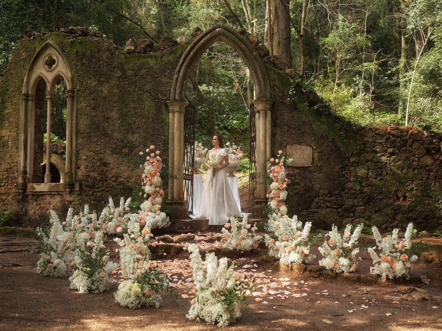 A romantic wedding ceremony setup in ancient stone ruins with floral arrangements and rose petals scattered on the ground.