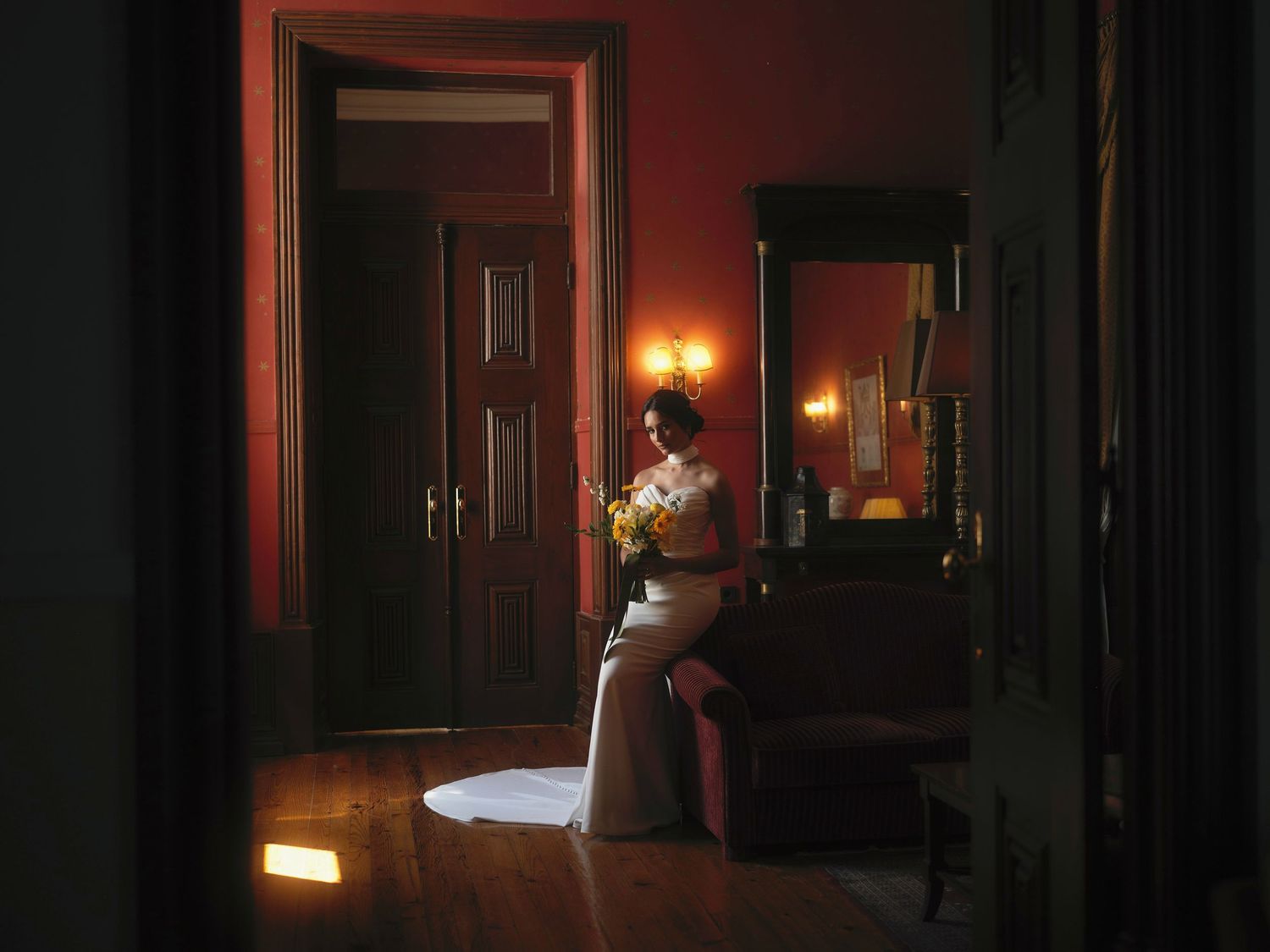 Bride in elegant gown holding bouquet stands in dimly lit Victorian room with warm red walls and antique mirror.