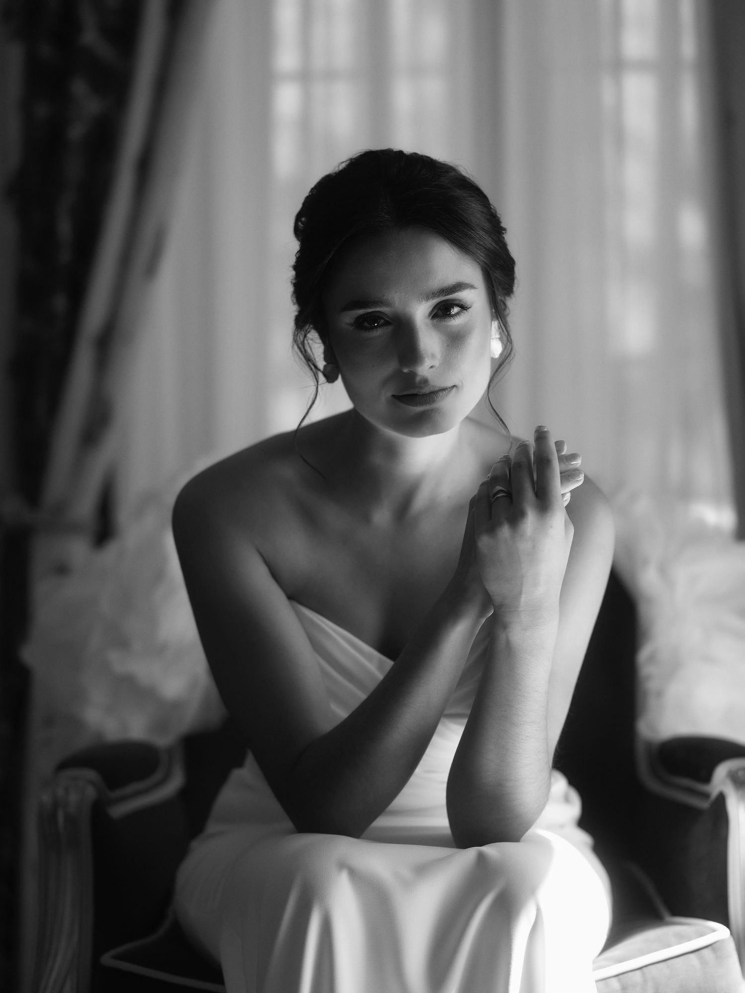 Black and white portrait of an elegant bride in a strapless gown, seated gracefully near a window.
