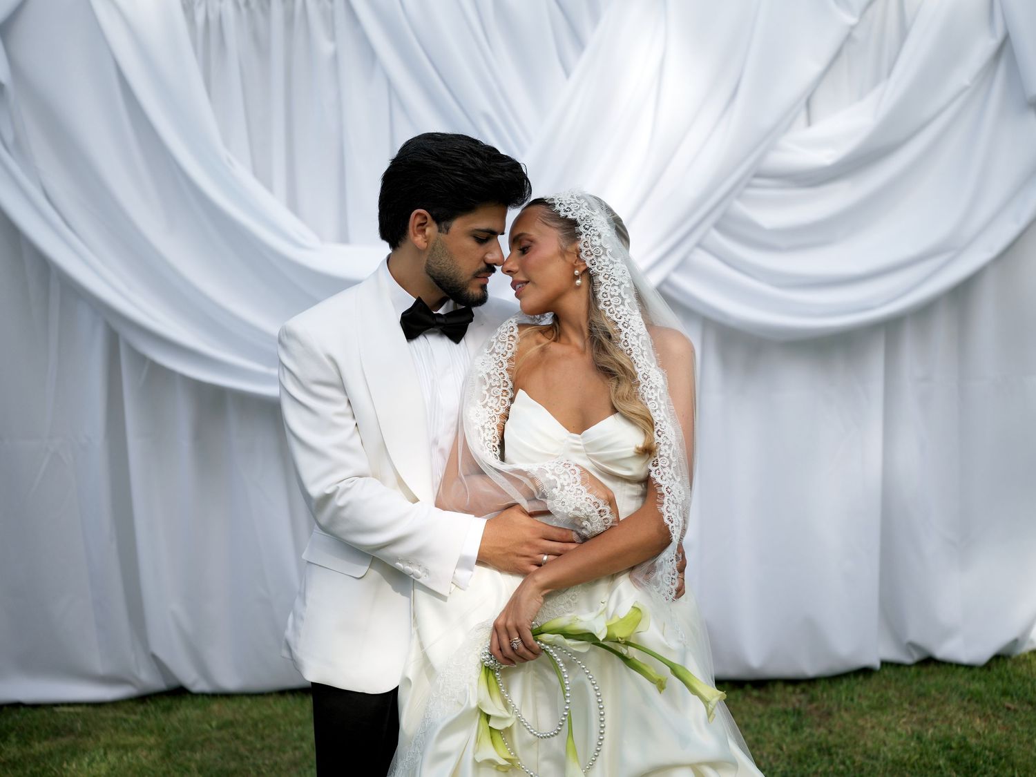 Elegant bride and groom in white wedding attire pose romantically before draped white fabric backdrop outdoors.