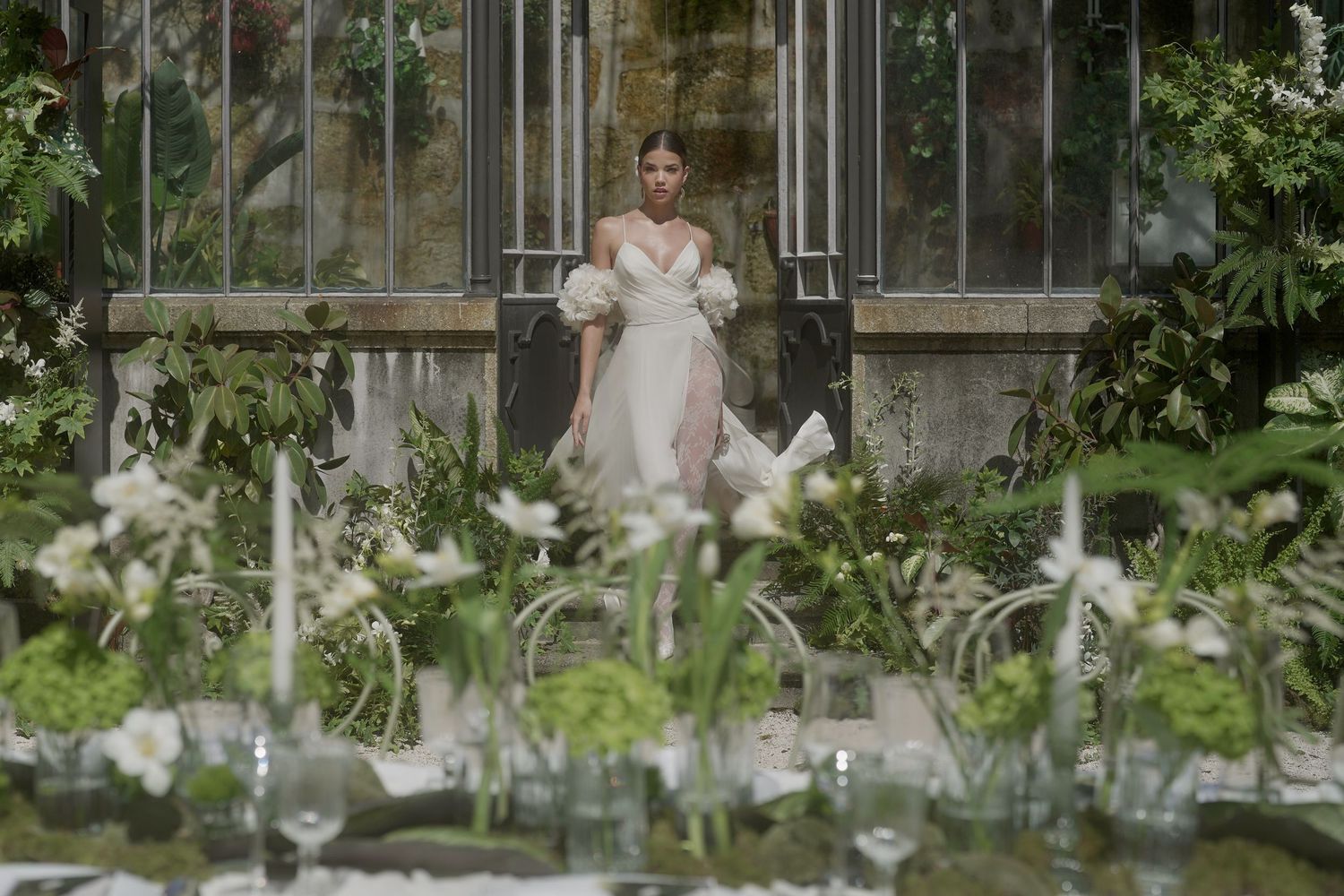 A person in a white dress poses elegantly in a greenhouse setting surrounded by blooming white lilies and greenery.