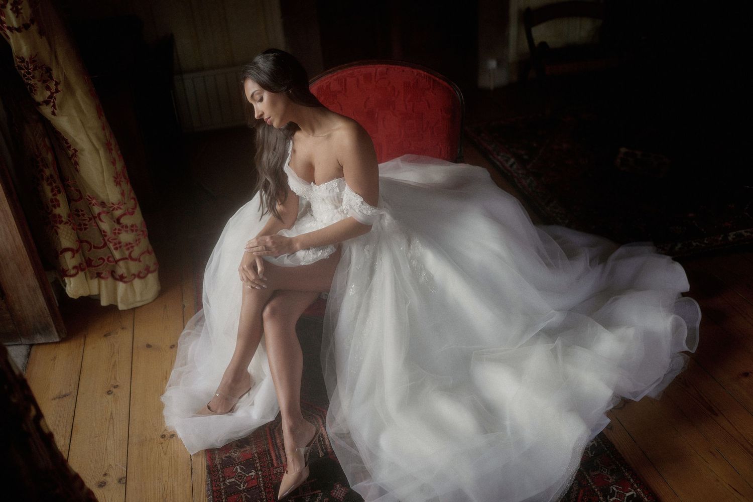 A bride in a flowing white wedding gown sits elegantly on a rustic wooden trunk in a dimly lit room.