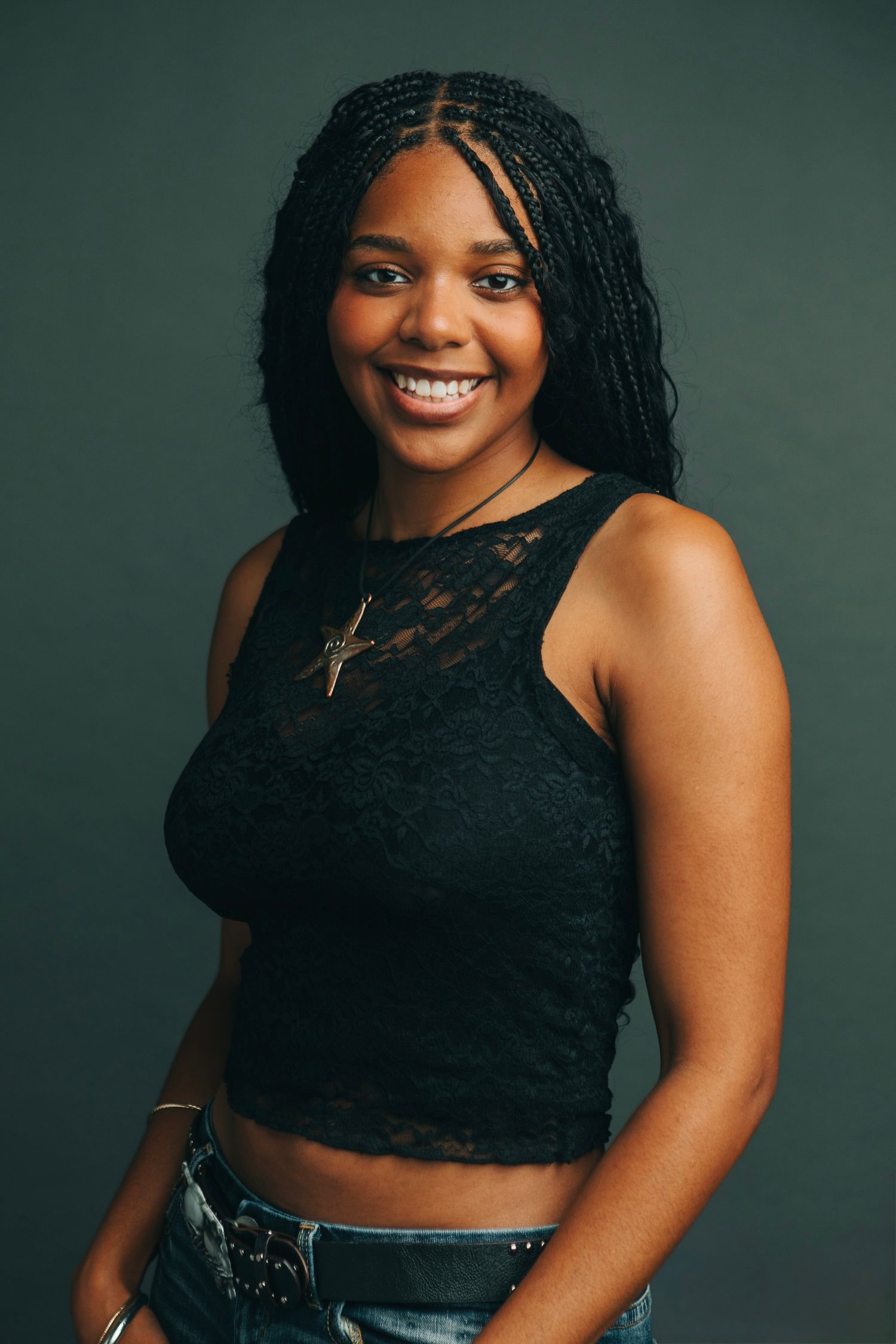 Young Black woman with box braids smiling, wearing black lace crop top and cross necklace against dark background.
