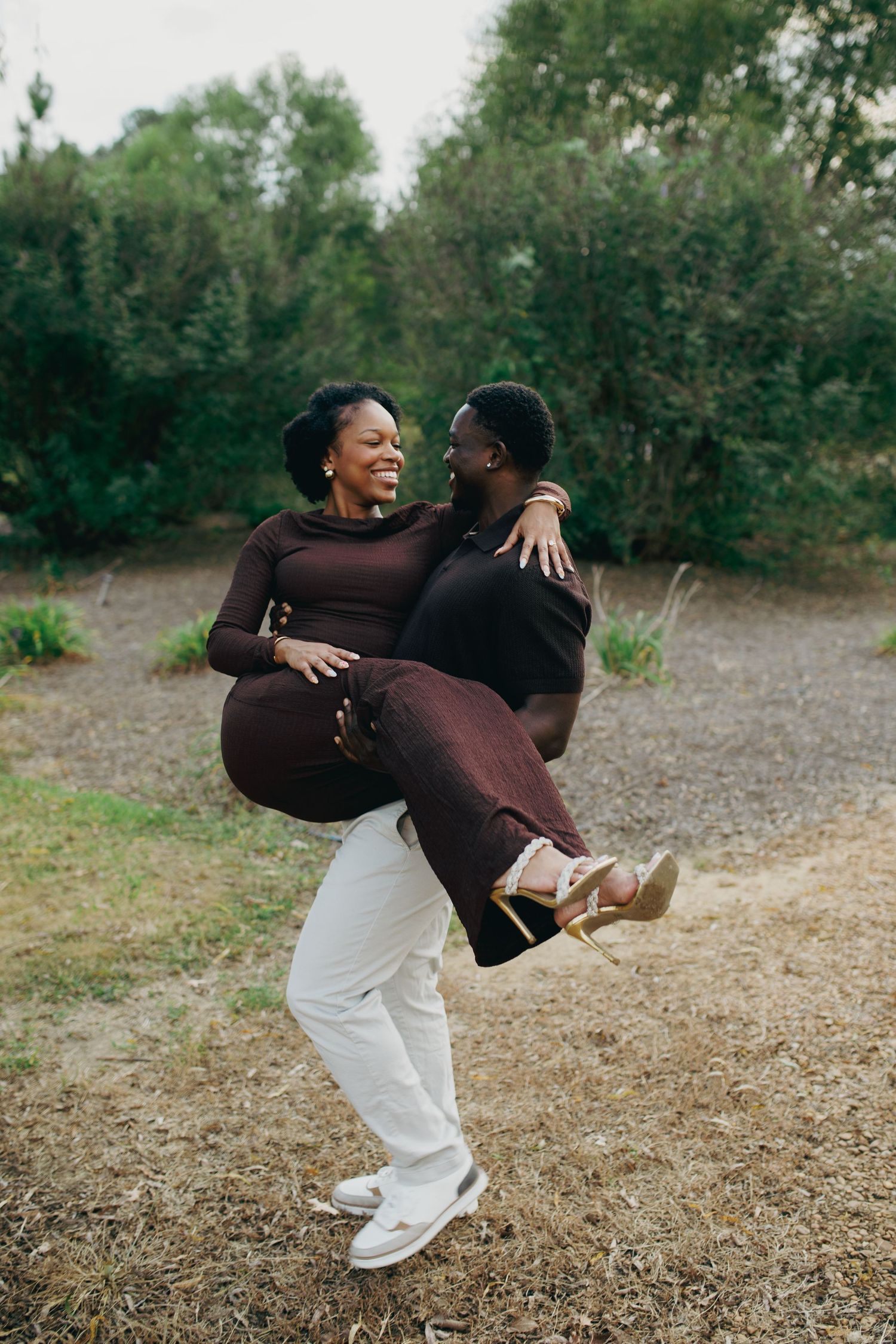 A man joyfully carries a laughing woman in his arms outdoors, both dressed in coordinated brown and white outfits.