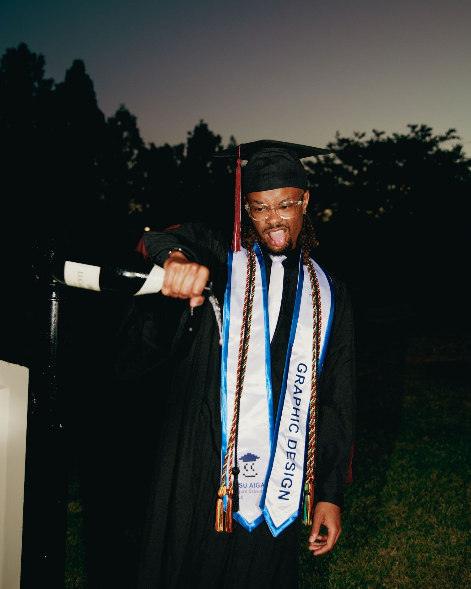 Graduate in black cap and gown with Graphic Design stole celebrates holding diploma outdoors at dusk.