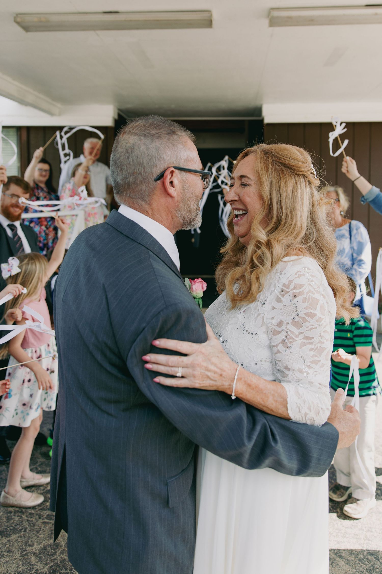 A joyful bride in lace dress embraces groom in gray suit as wedding guests celebrate with ribbon wands outdoors.