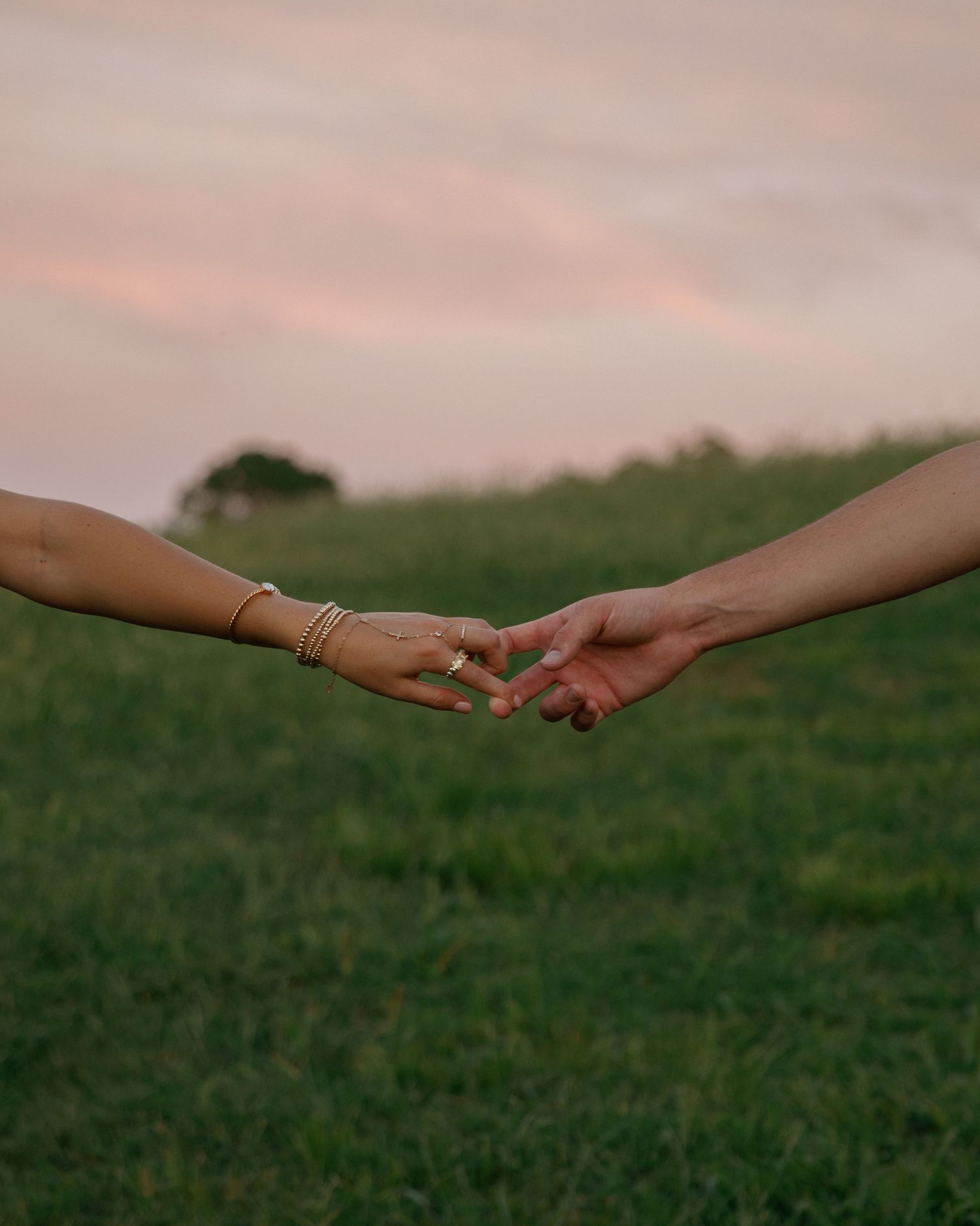 Two hands gently touching fingertips in a green field at sunset, one adorned with gold bracelets and a ring.