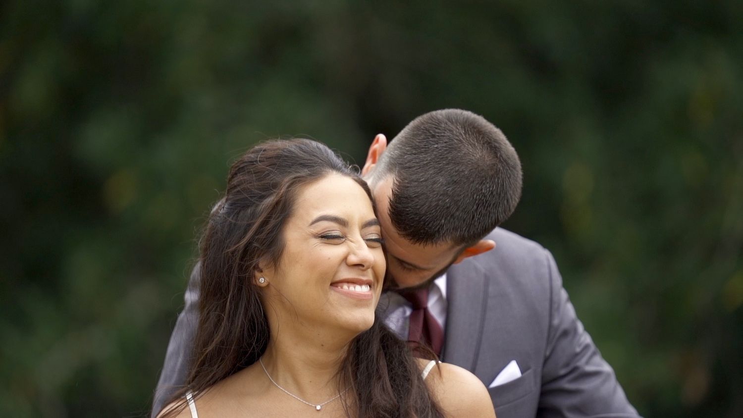 A joyful wedding couple share an intimate moment outdoors with lush greenery in the background.