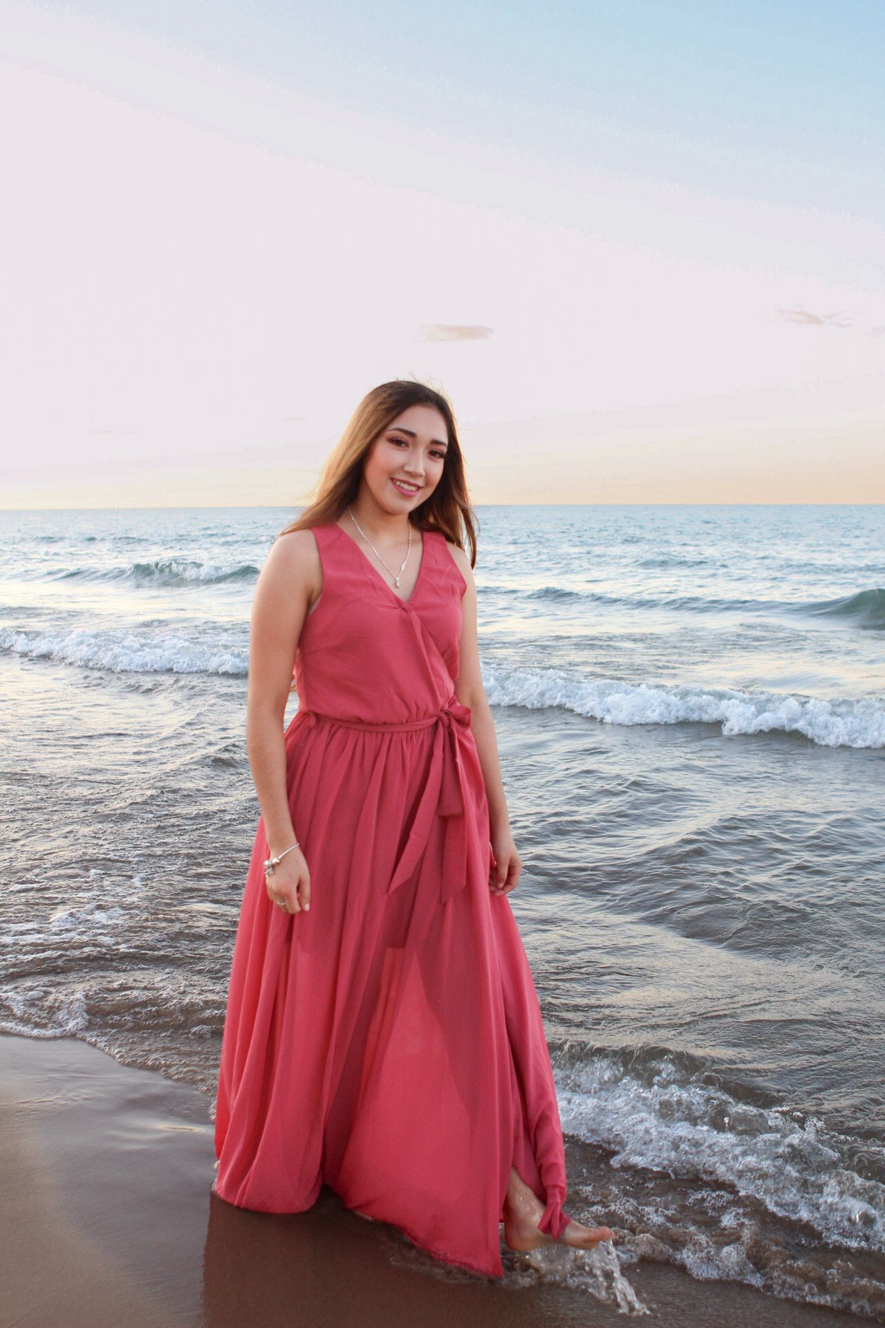 Person in coral dress poses playfully at beach with waves in background.