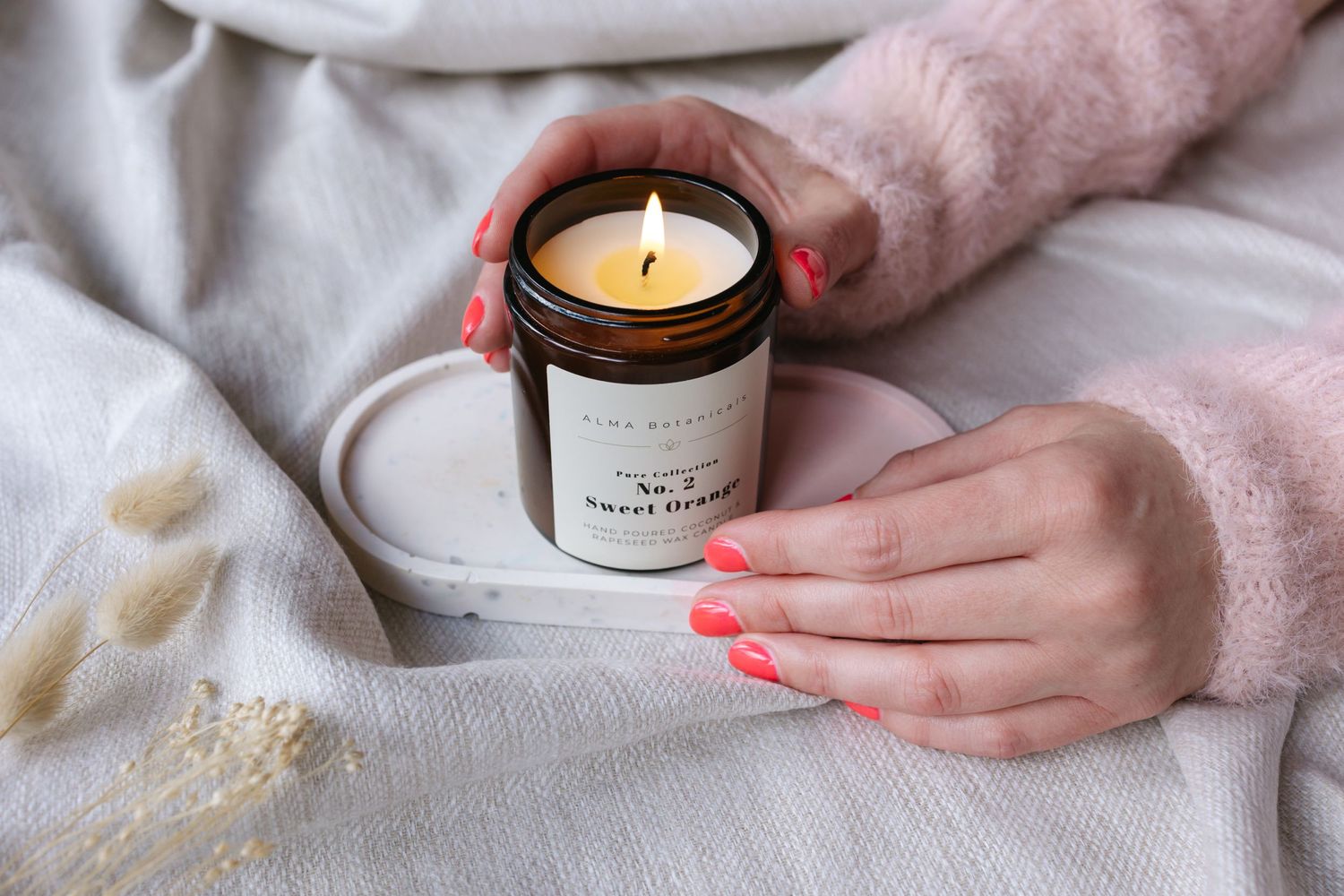 Hands with coral pink nails hold a minimalist scented soy candle against soft white and pink textiles.