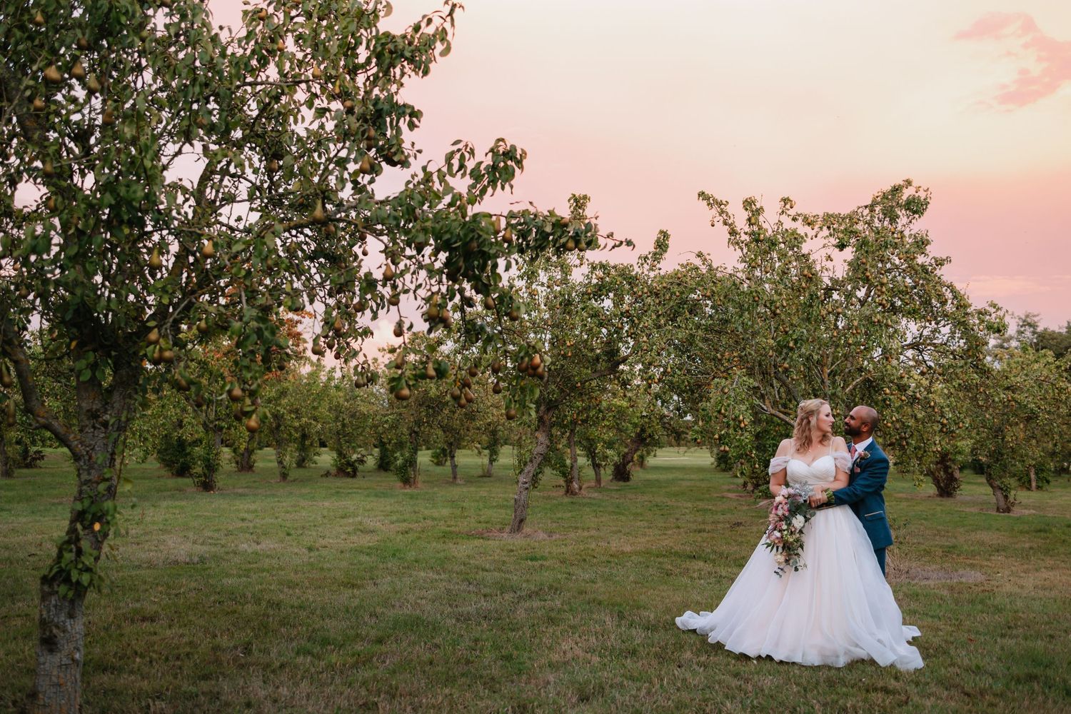 A couple embraces in a tranquil orchard during golden hour with pink sunset skies and flowering trees.

Prestwold Hall