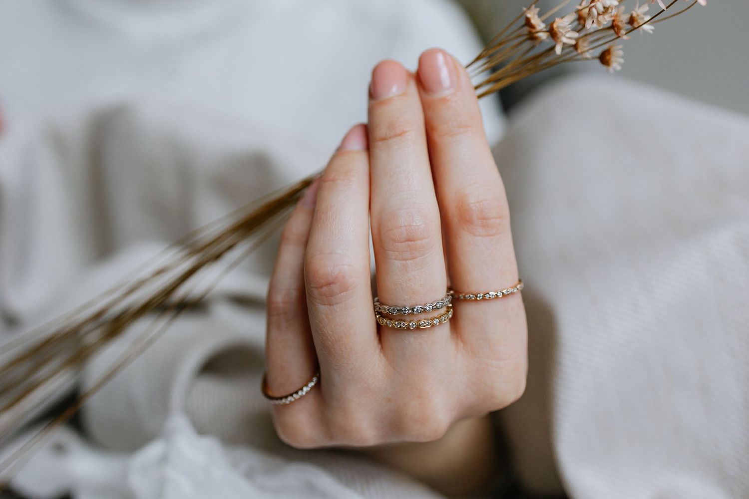 Delicate hand wearing multiple dainty gold rings against white fabric with dried flowers.