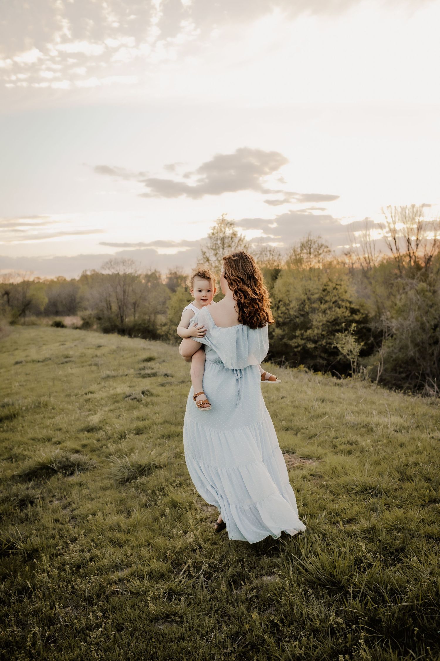 Mom and baby walking through the field at sunset. Dress blowing in the wind.  Captured in St. Charles County. Gorgeous sky.