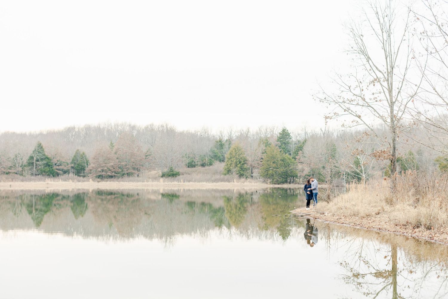 A person stands at the edge of a serene lake surrounded by bare winter trees and evergreens on a misty morning.