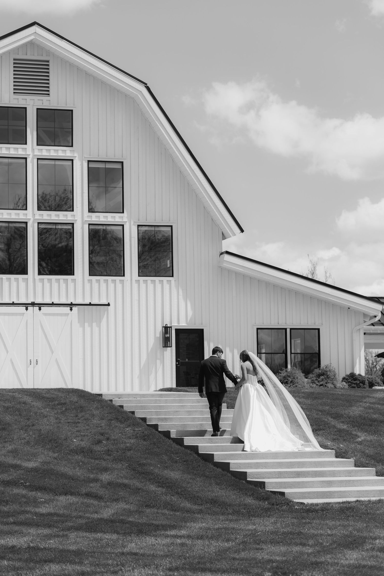 Bride and groom walking up steps toward white barn wedding venue, black and white photo.