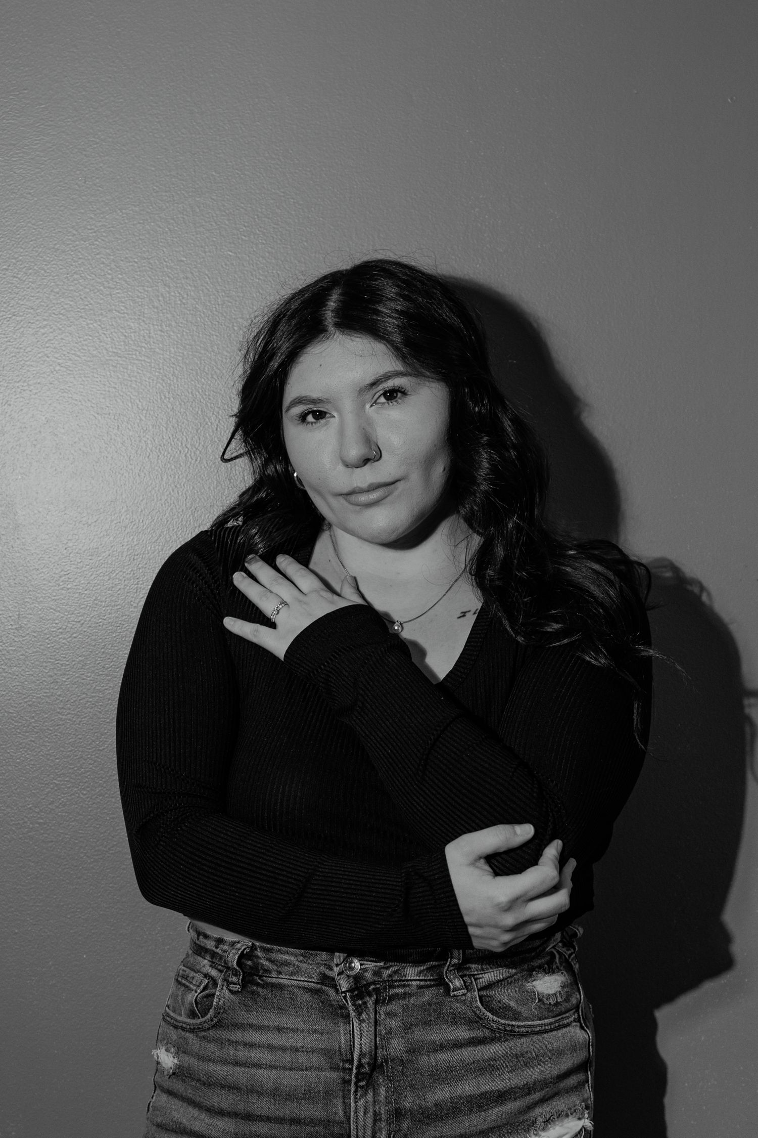 Young woman in black top and jeans posing confidently against wall in black and white portrait.