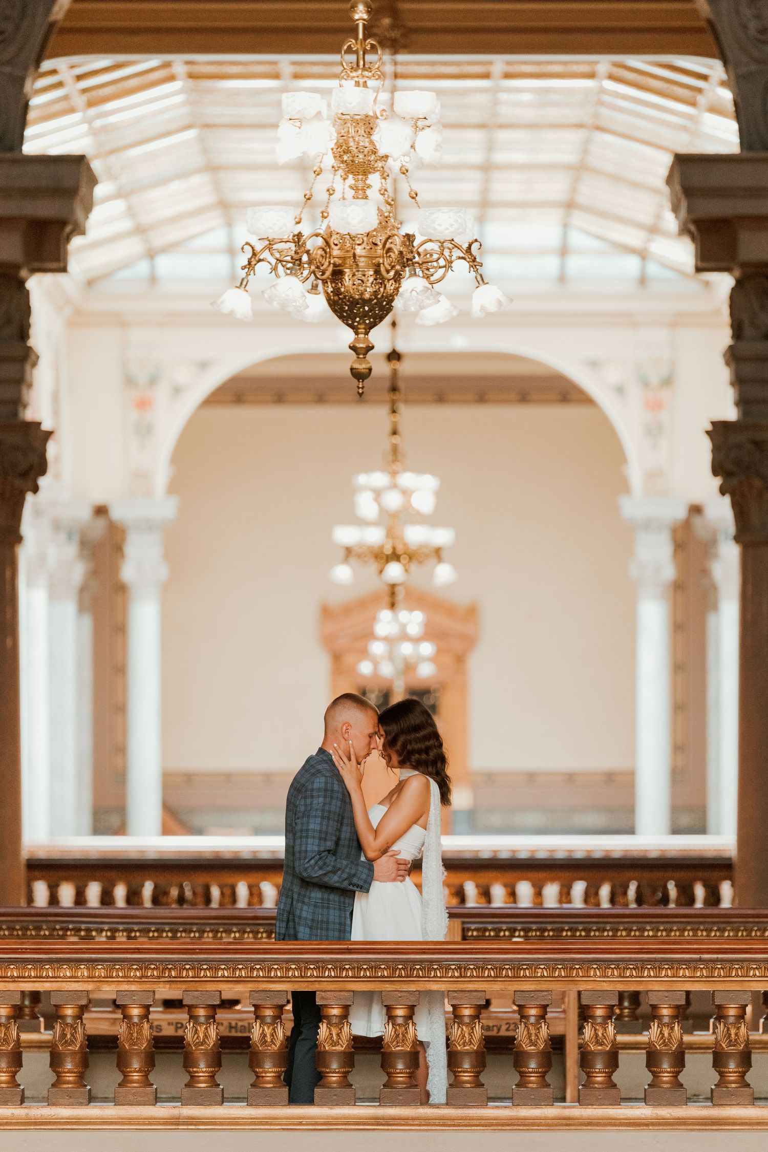 Couple sharing romantic kiss on ornate balcony beneath grand chandelier in elegant historic venue.