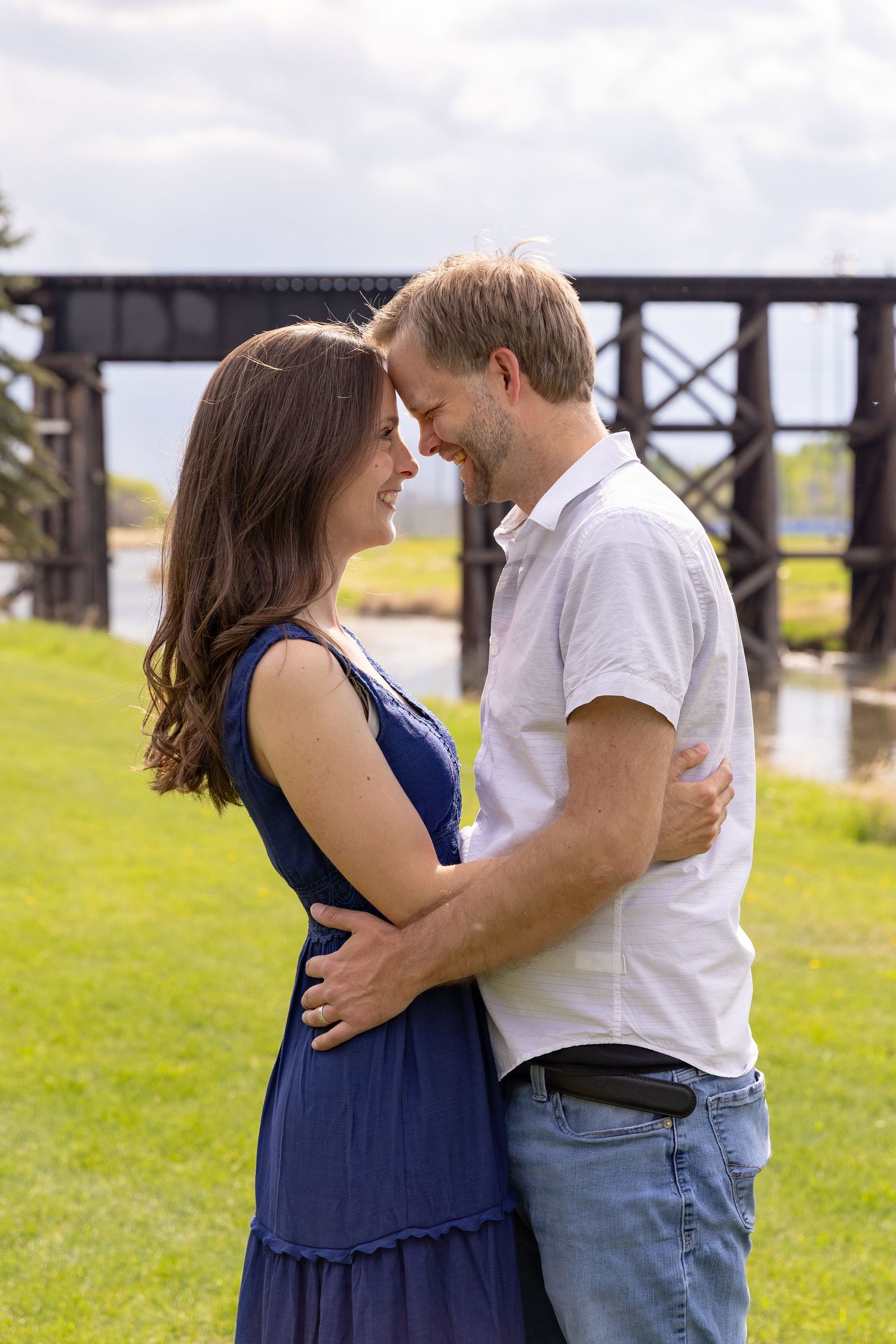 Smiling couple embracing face-to-face on green grass with a wooden trestle bridge behind them.