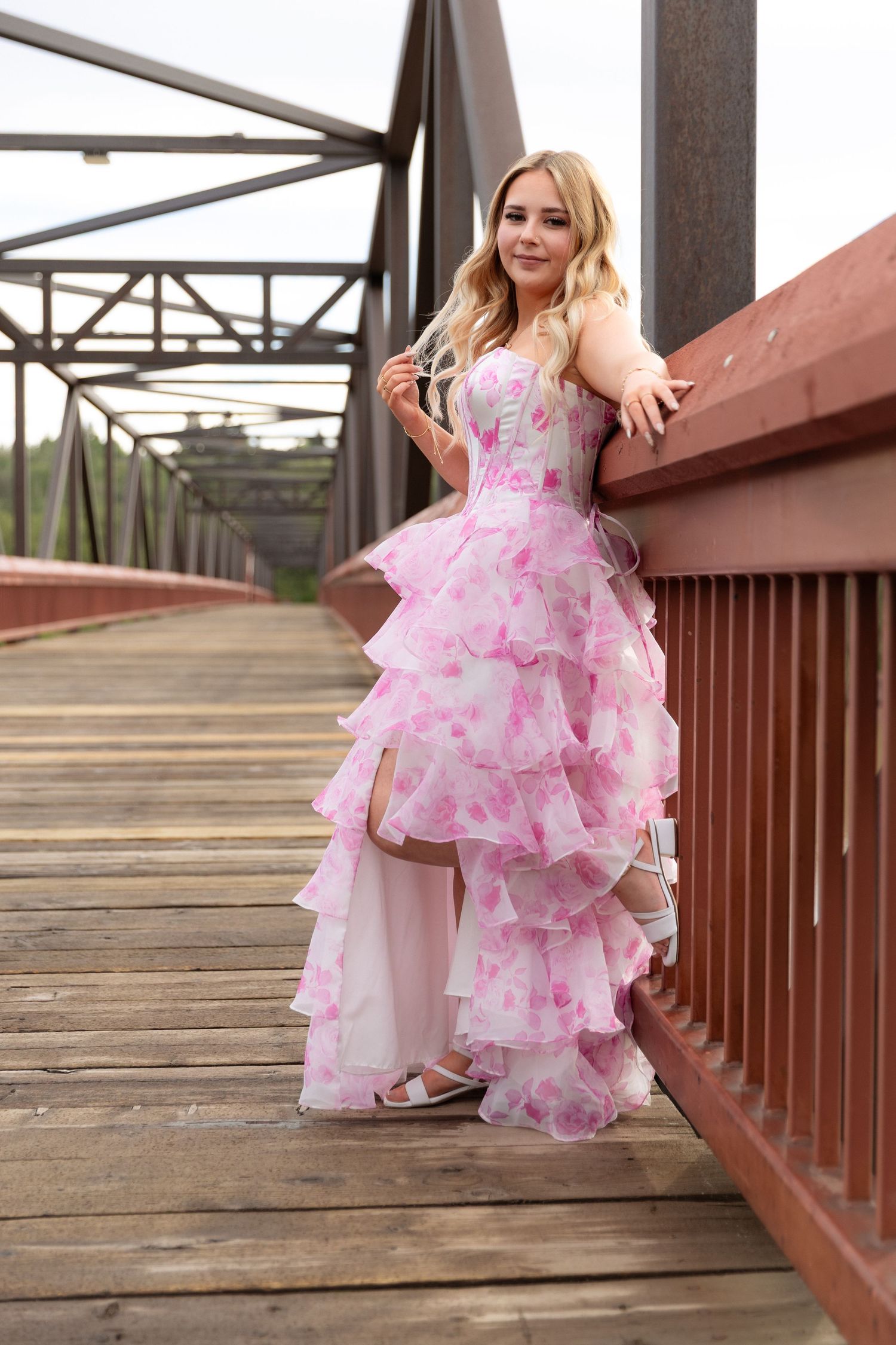 Blonde teen in pink layered floral prom gown standing on wooden bridge railing.