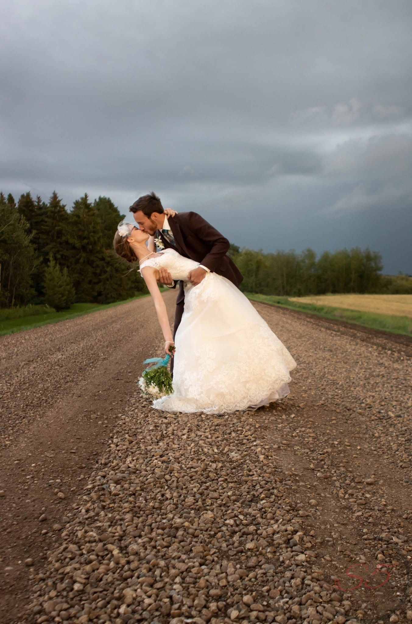 Bride and groom sharing a romantic dip kiss on a gravel country road under dramatic stormy skies.