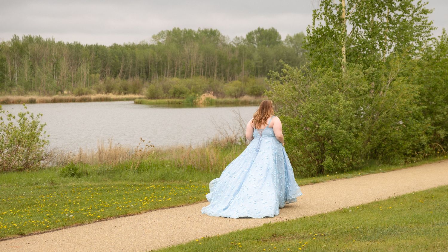Young woman in a flowing light blue ball gown walking along a path near a serene lake surrounded by green trees.