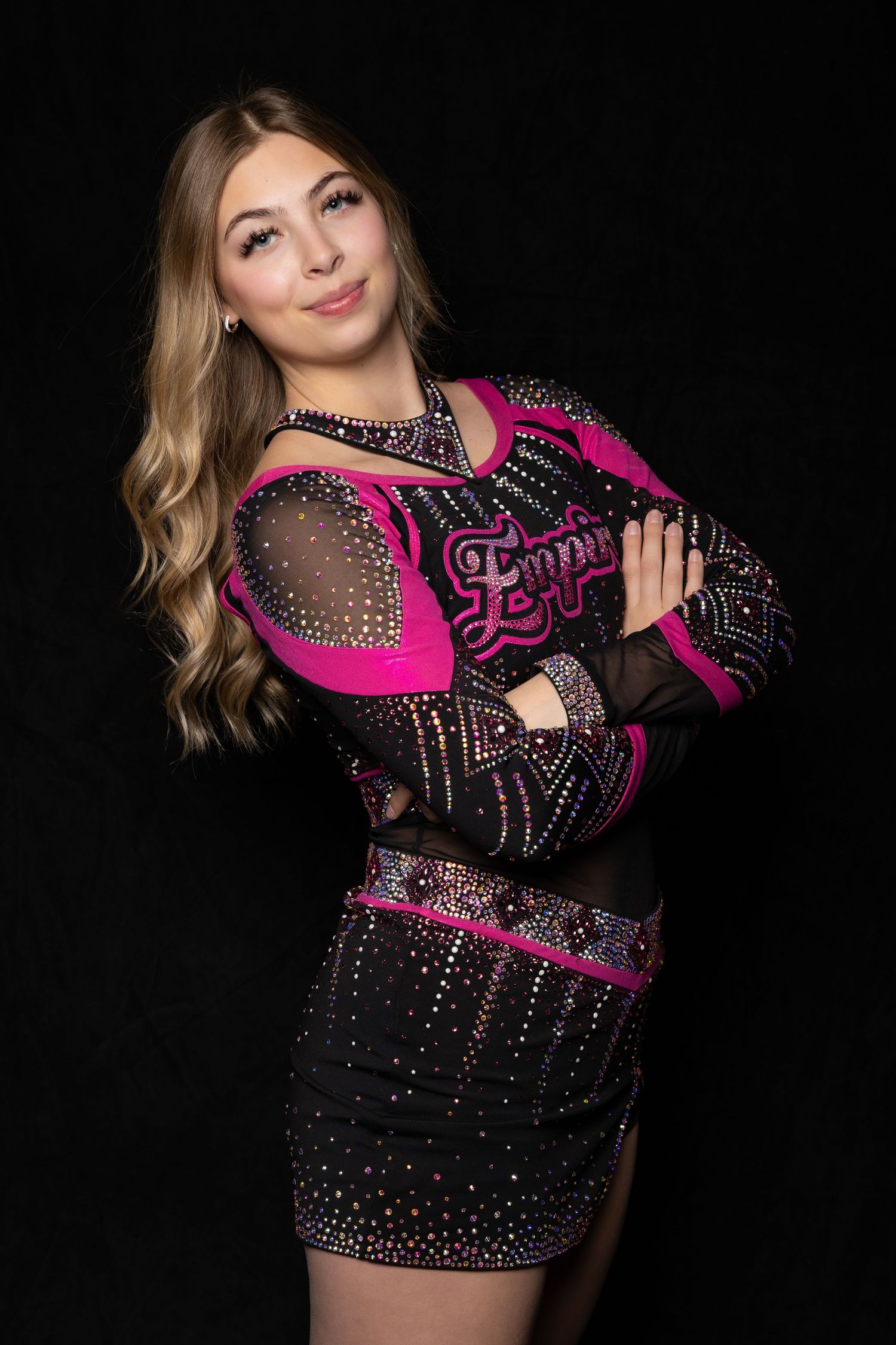 Blonde girl in black and pink rhinestone Empire cheer uniform with arms crossed.