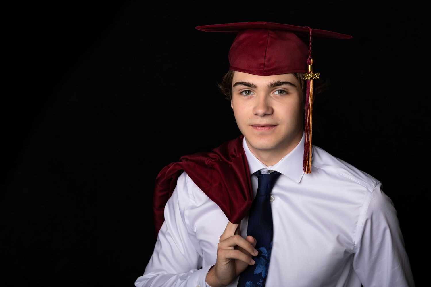 Teen boy in graduation cap and gown smiling against dark studio background.