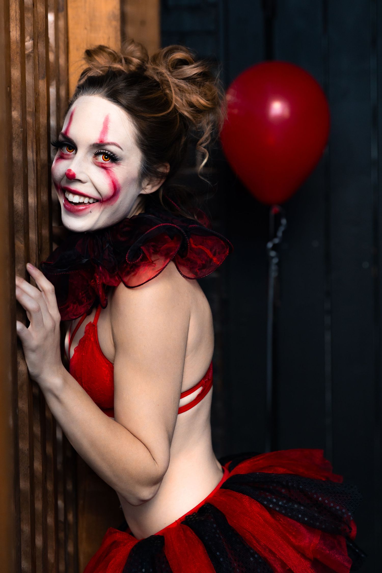 Woman in red clown costume with face paint laughing while peeking around a wooden door with red balloon.