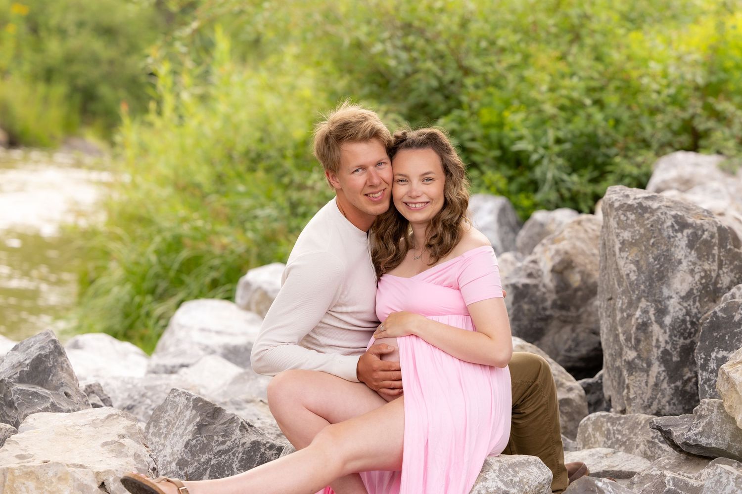 Pregnant couple sitting on rocks outdoors, man embracing woman in pink maternity dress, smiling.