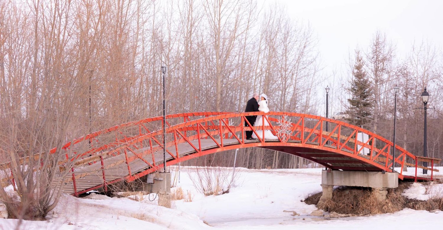 A couple shares a romantic kiss on a red arched bridge in a snowy landscape.