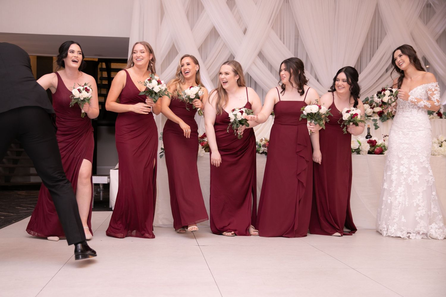 Bridesmaids in burgundy gowns laughing and walking with bouquets alongside bride at wedding reception.