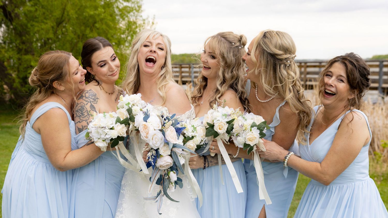 Bride in white gown laughing joyfully with bridesmaids wearing light blue dresses holding white bouquets.