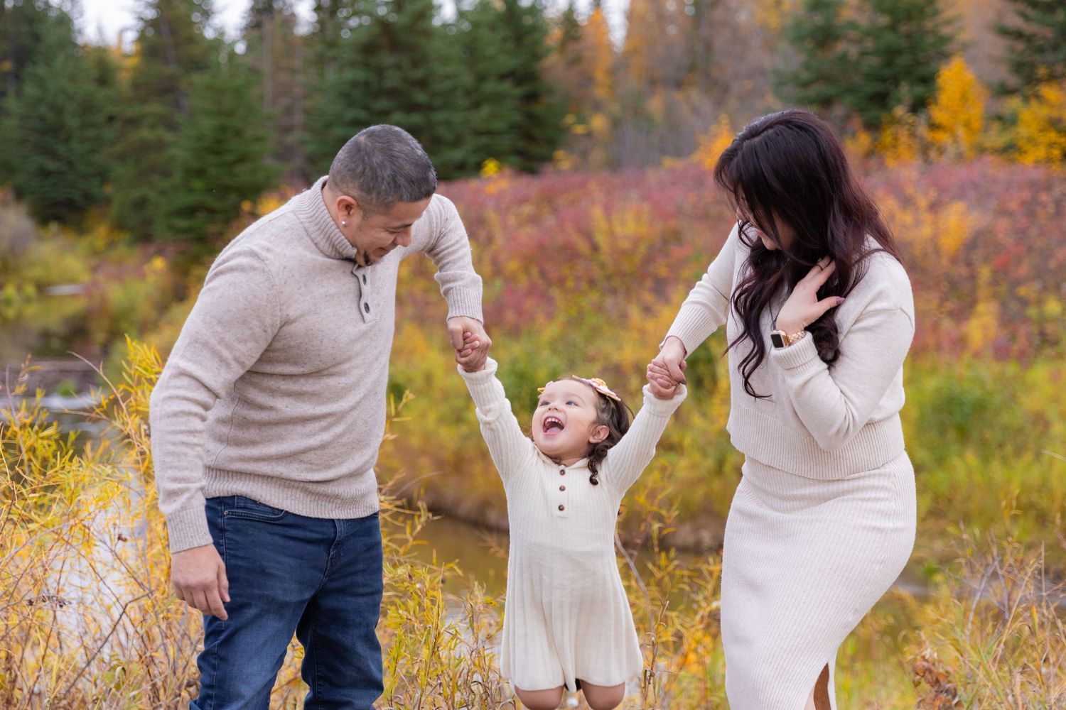 Parents joyfully swing their toddler daughter by hands in colorful autumn outdoor setting.