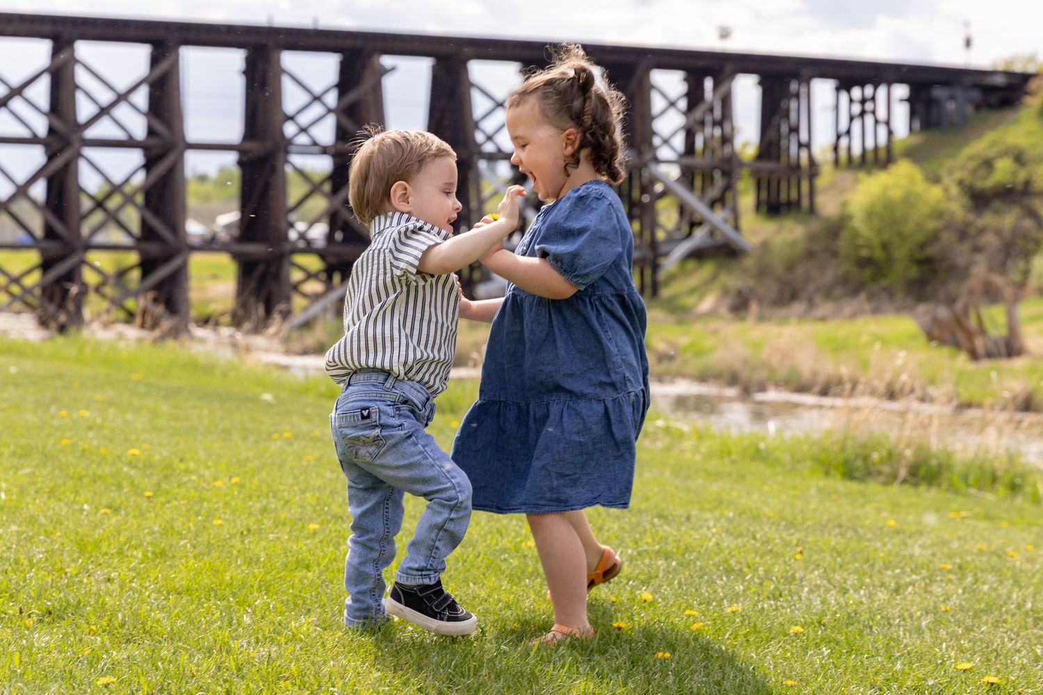 Two young children playfully dancing together on green grass near a wooden bridge structure.