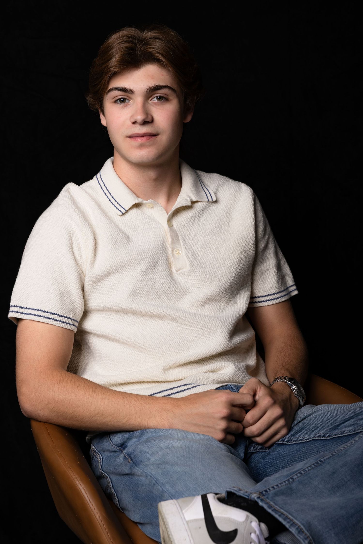 Teen boy in white polo shirt sitting relaxed in chair against black background.