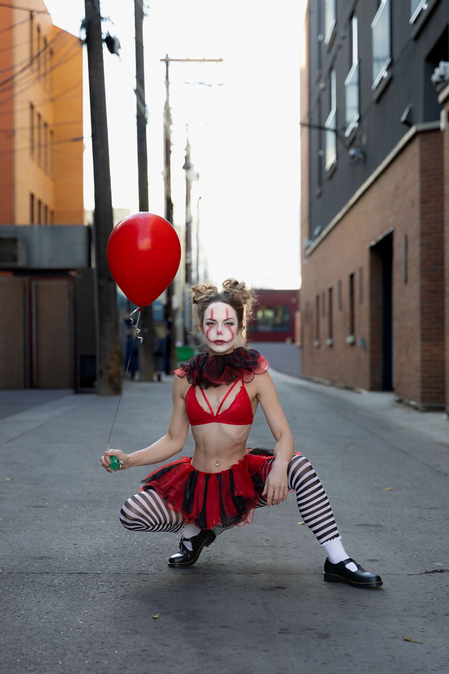 Female clown in red costume with striped stockings crouching in an urban alley holding a red balloon.