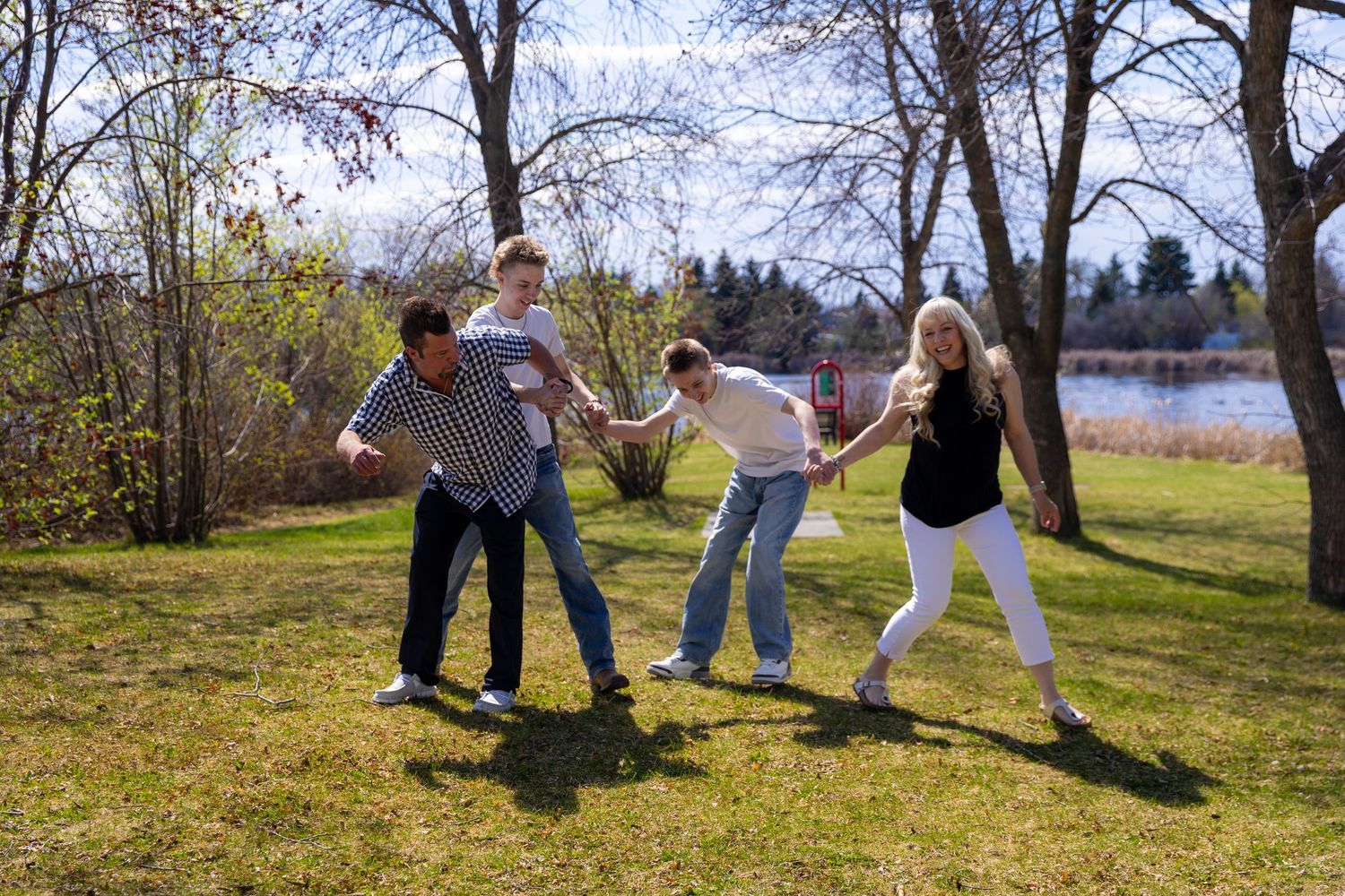 Playful family of four laughing and pulling each other in a grassy park near a lake in spring.