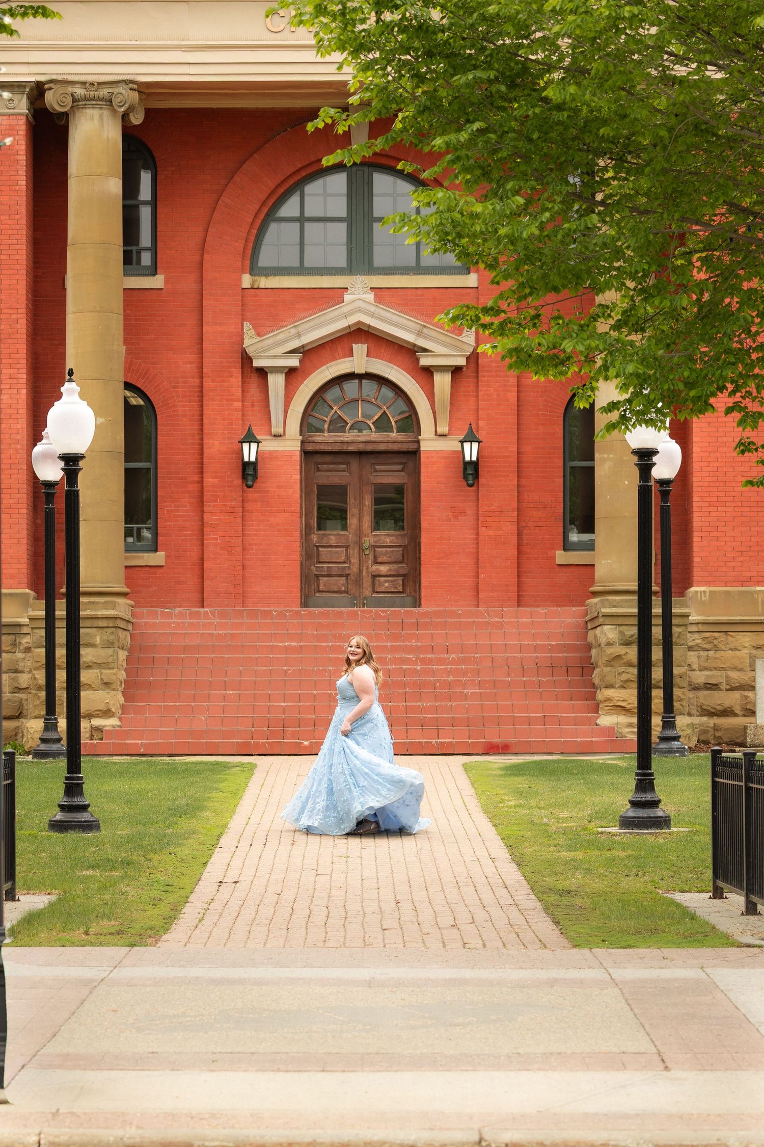 Woman in blue gown walking on brick path before a grand red brick building.