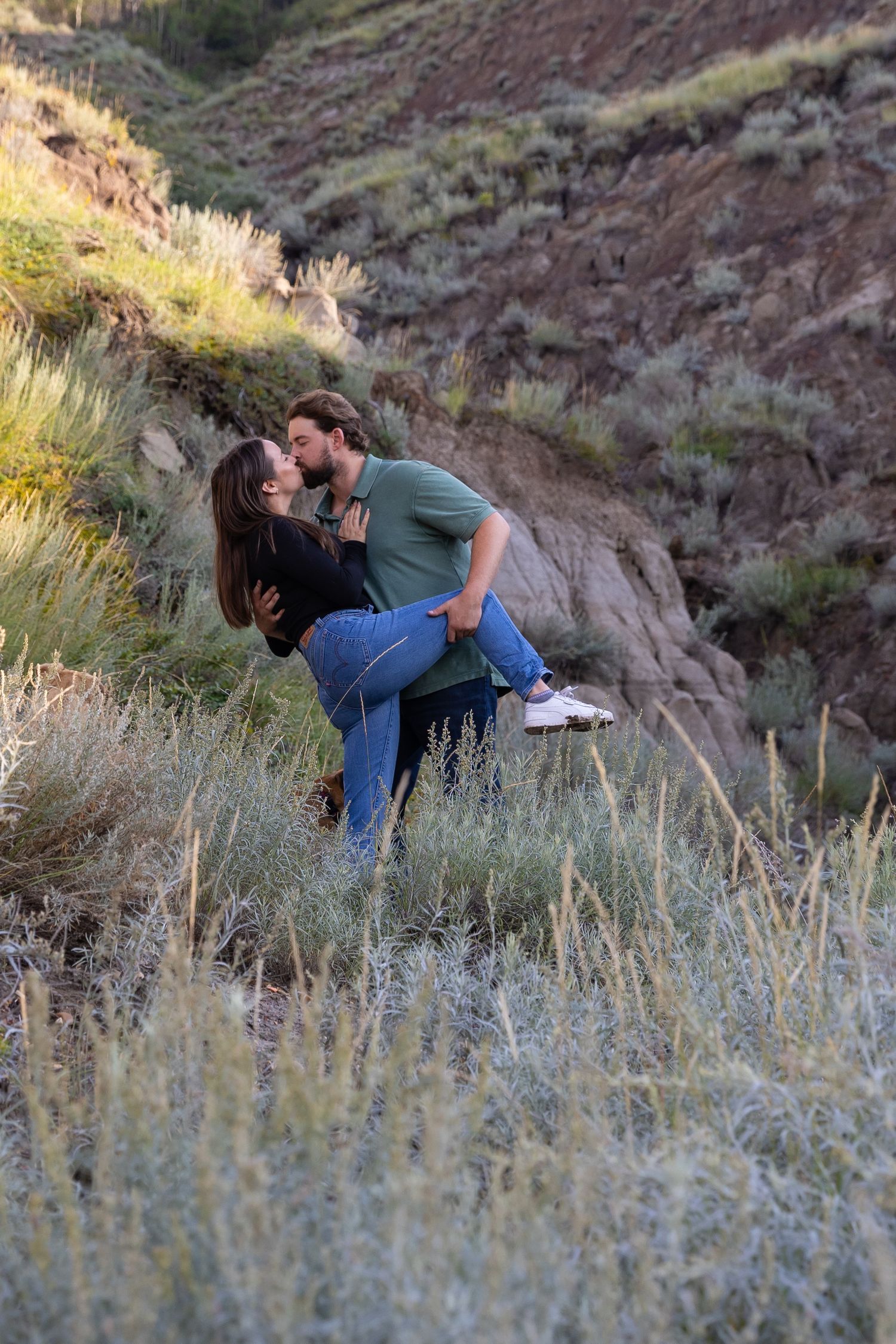 Man lifting and kissing woman in a romantic outdoor setting among wild grass and hills.