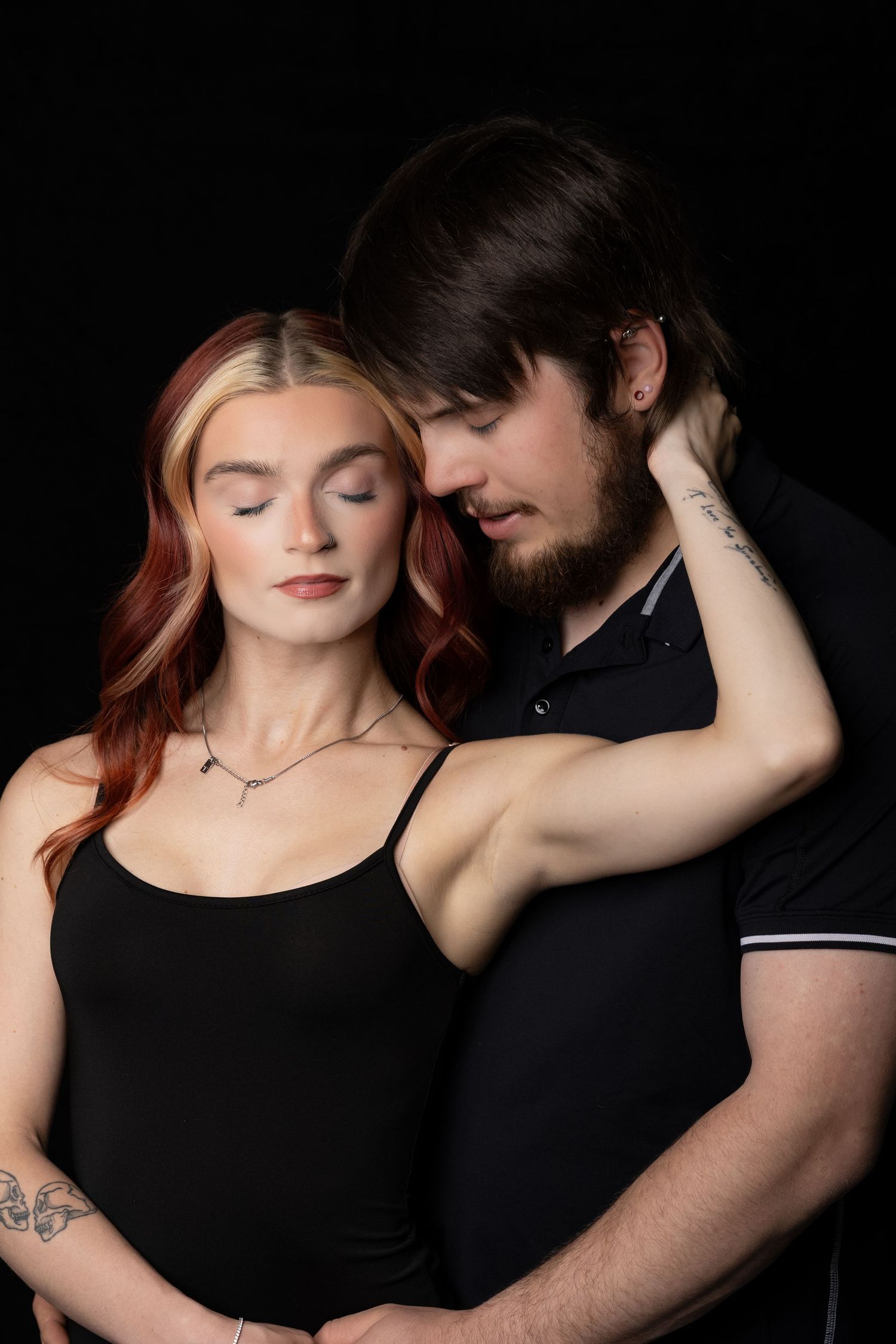 A romantic couple in black outfits share a tender intimate moment together against a dark studio background.