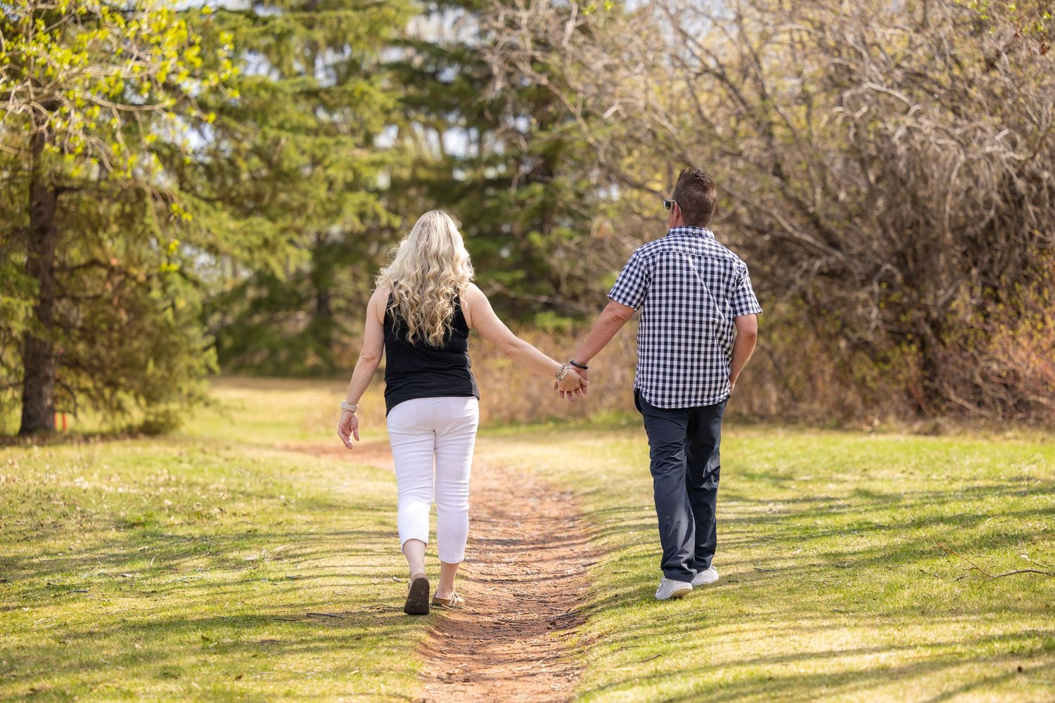 Couple holding hands walking down a scenic park path surrounded by lush green grass and autumn trees.
