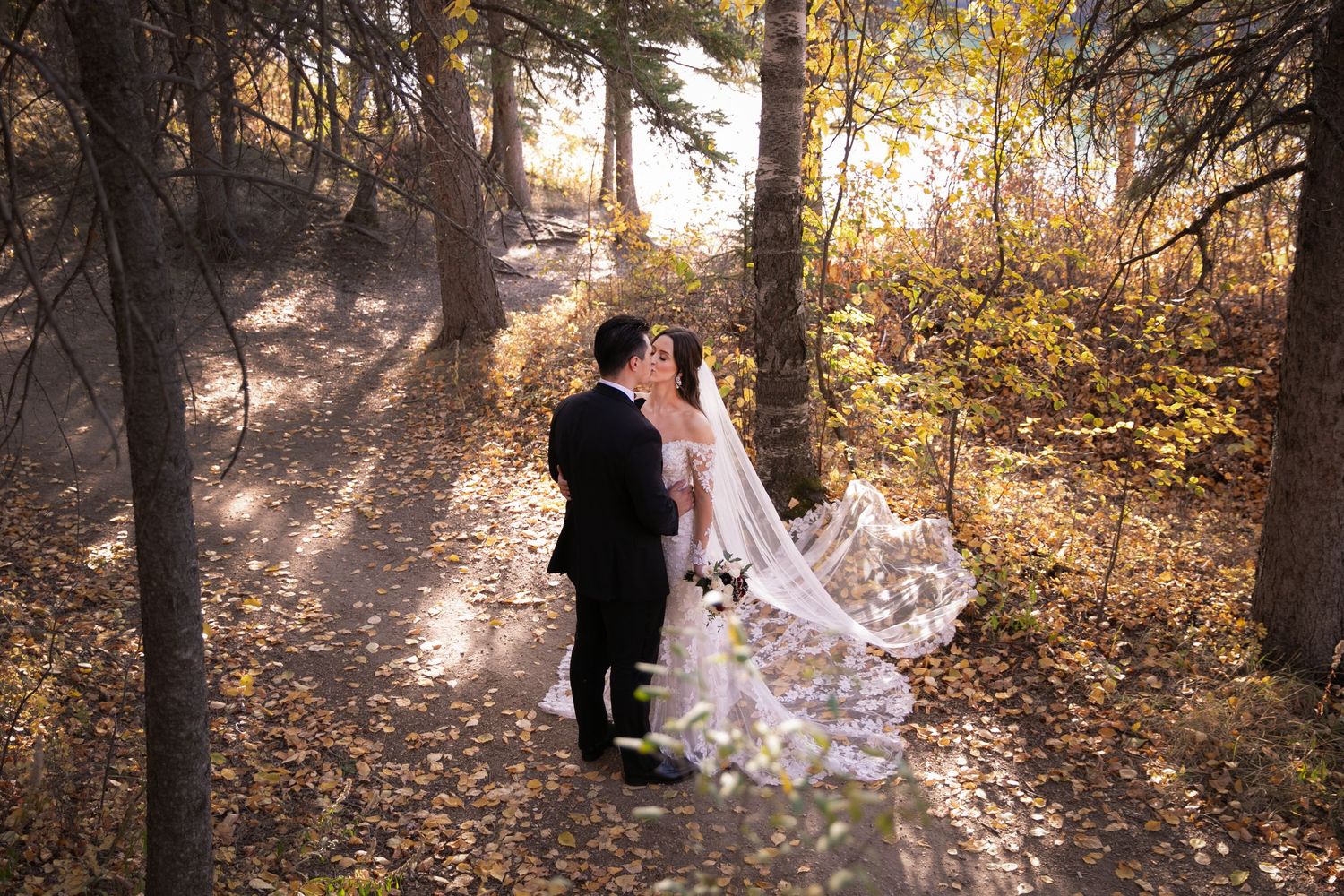 Bride and groom sharing a romantic kiss on a forest path surrounded by autumn foliage with veil flowing in the breeze.