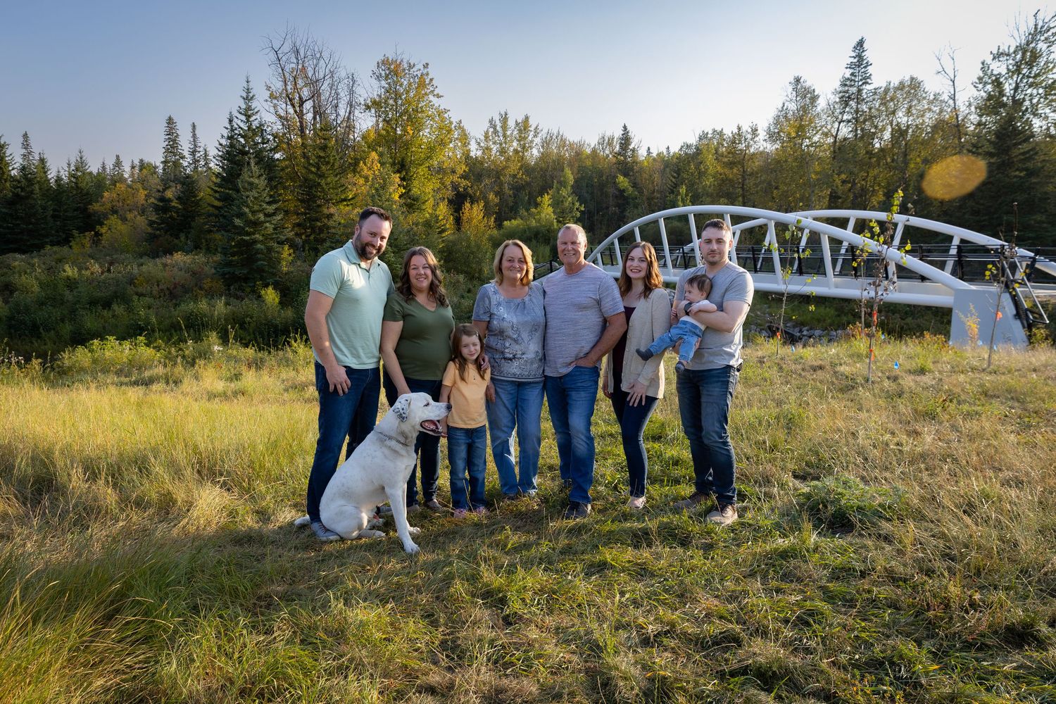 Family of six with a white dog posing outdoors near a white bridge surrounded by green trees and meadow.