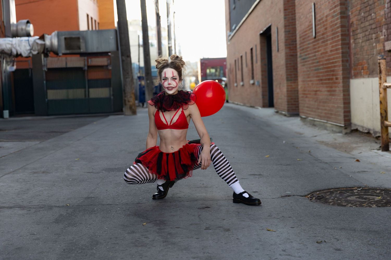 A woman in clown costume crouches in an alley holding a red balloon.