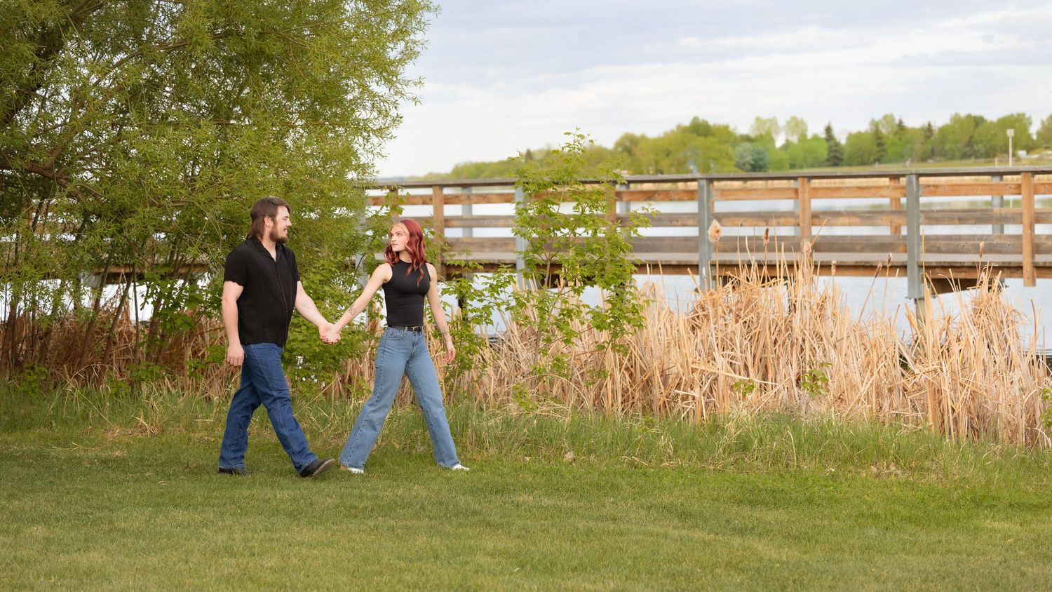 A couple walks hand-in-hand on green grass near a wooden bridge surrounded by tall reeds and trees. in Leduc Alberta