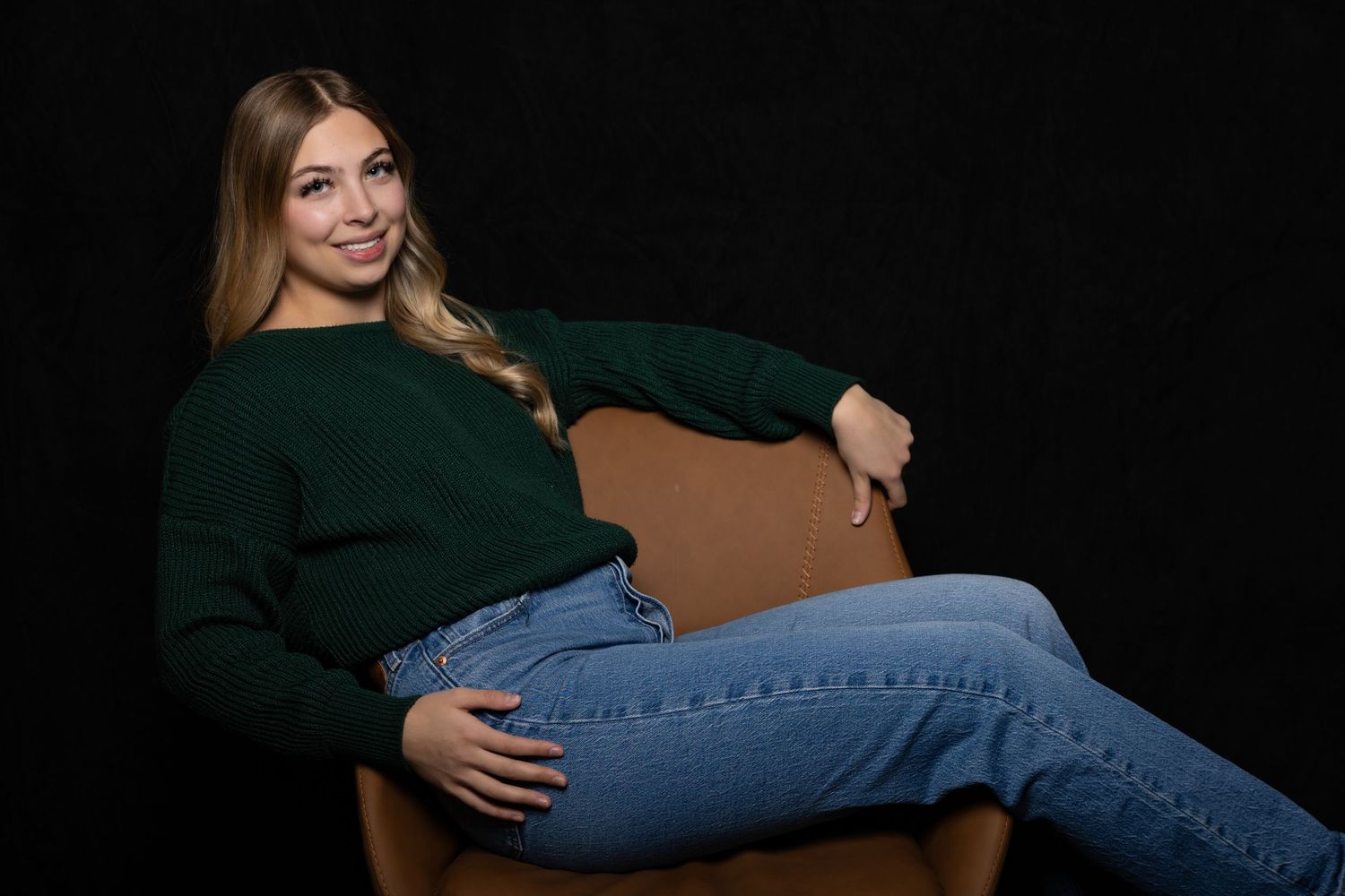 Smiling blonde woman in green sweater and jeans relaxing in brown leather chair.